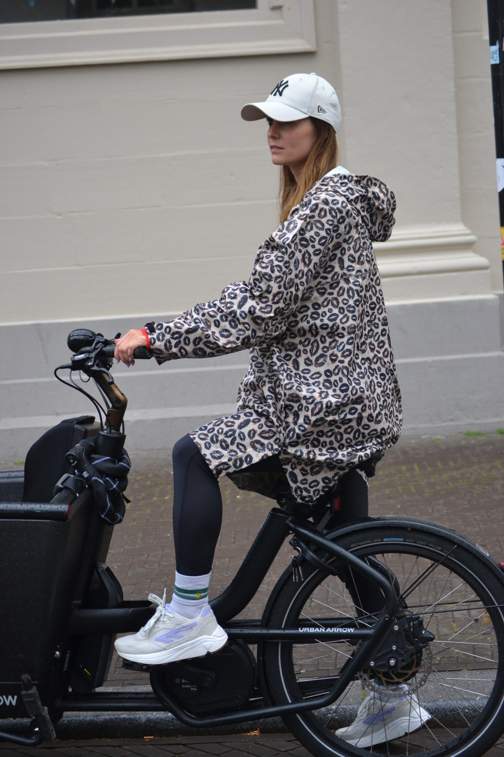 Vrouw fietst op een bakfiets in de stad terwijl ze een leopard regenponcho met capuchon draagt. Stijlvolle en praktische regenkleding voor dames.